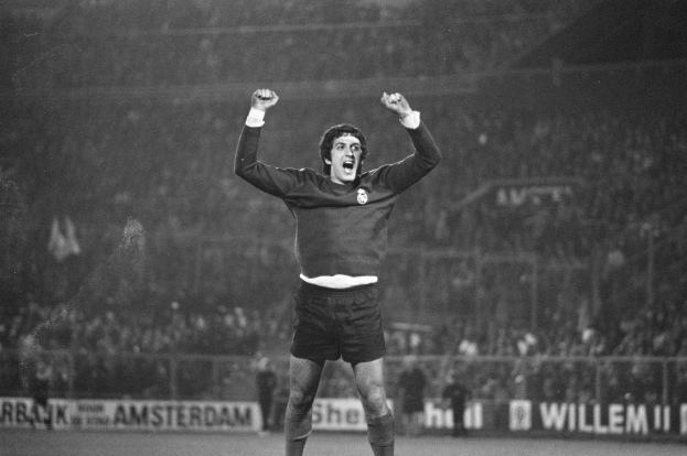 Ein Schwarz-Weiß-Foto eines Mannes, der auf einem Fußballfeld ein Tor feiert, mit erhobenen Armen und einem freudigen Gesichtsausdruck, umgeben von Menschen, einem Zaun und Schildern mit Text.