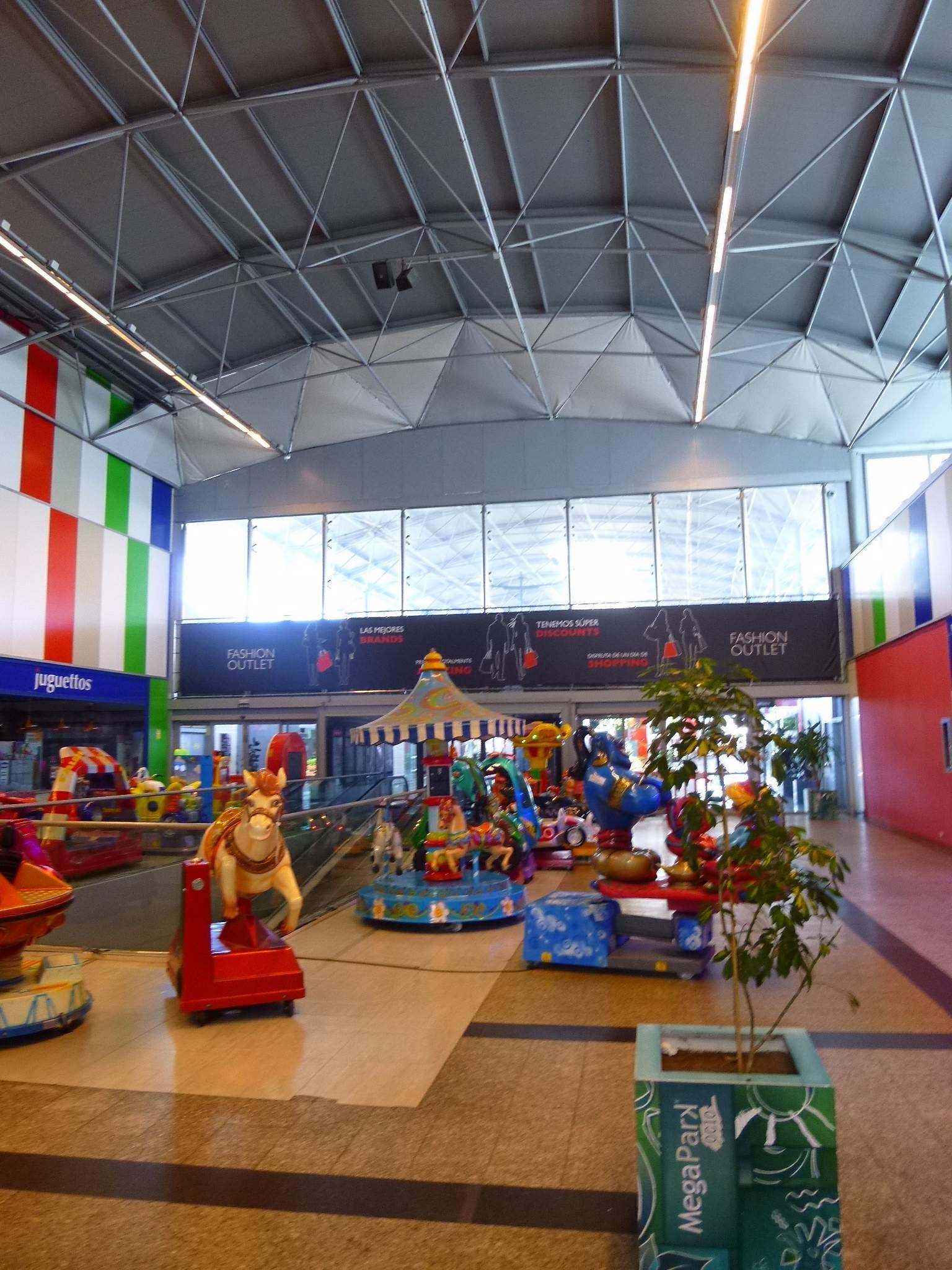 Großer Indoor-Vergnügungsbereich in einem Einkaufszentrum mit verschiedenen Fahrgeschäften, einem Topf mit einer Pflanze, verstreuten Spielzeugen, Informationsschildern, Deckenleuchten und Metallträgern.