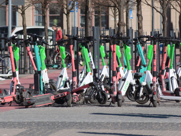 Eine Reihe von Elektro-Rollern, die auf einer Straße geparkt sind, mit einer Person im Vordergrund und Baumwipfeln und Gebäuden im Hintergrund.