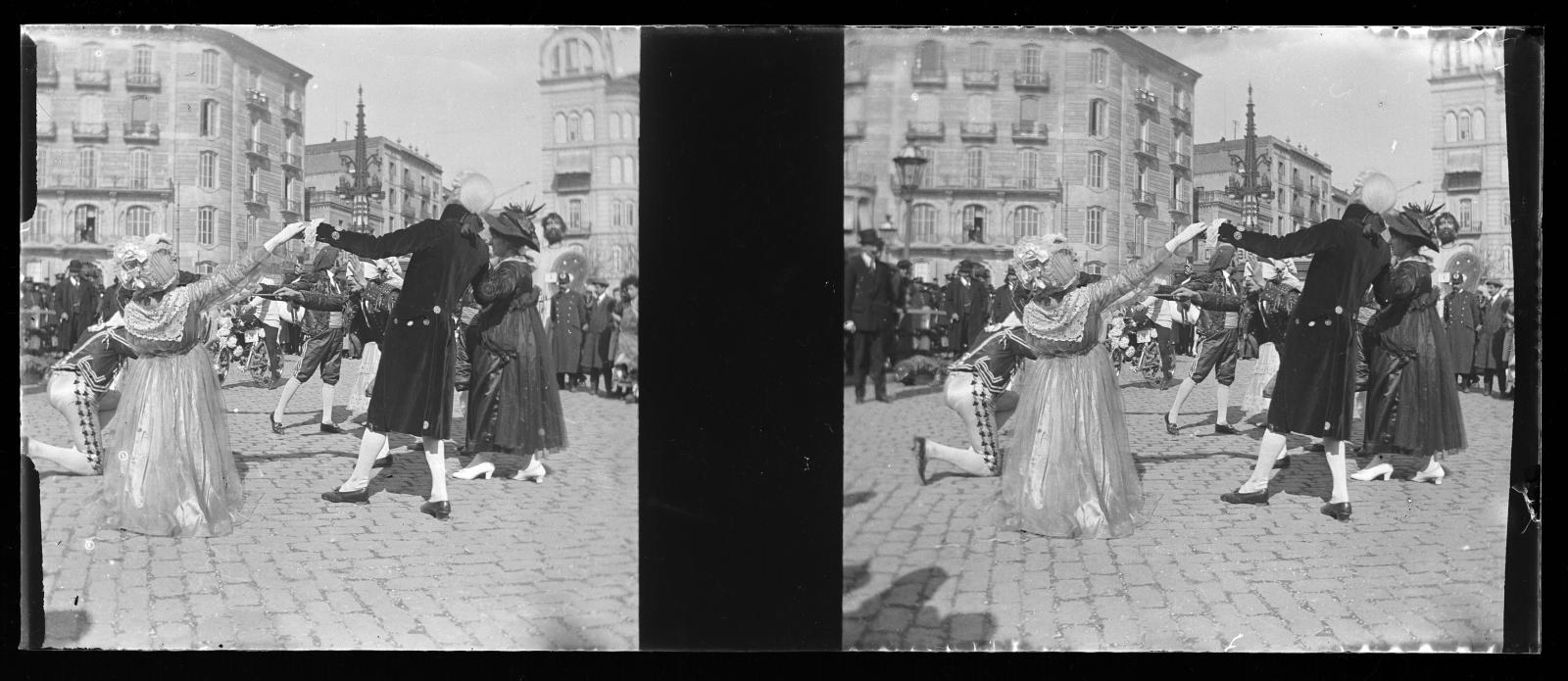 Schwarzes und weißes Foto von Menschen in Kostümen, die auf der Straße tanzen, umgeben von Gebäuden, Straßenlaternen und Lichtern unter einem klaren Himmel.
