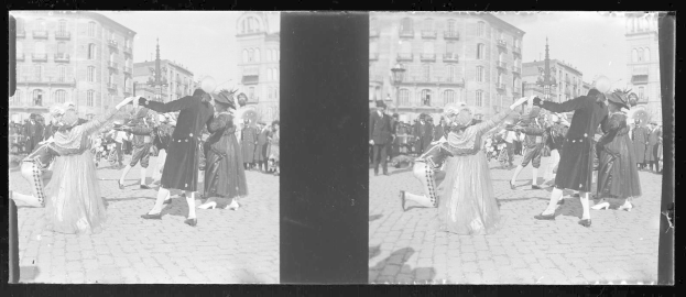 Schwarzes und weißes Foto von Menschen in Kostümen, die auf der Straße tanzen, umgeben von Gebäuden, Straßenlaternen und Lichtern unter einem klaren Himmel.
