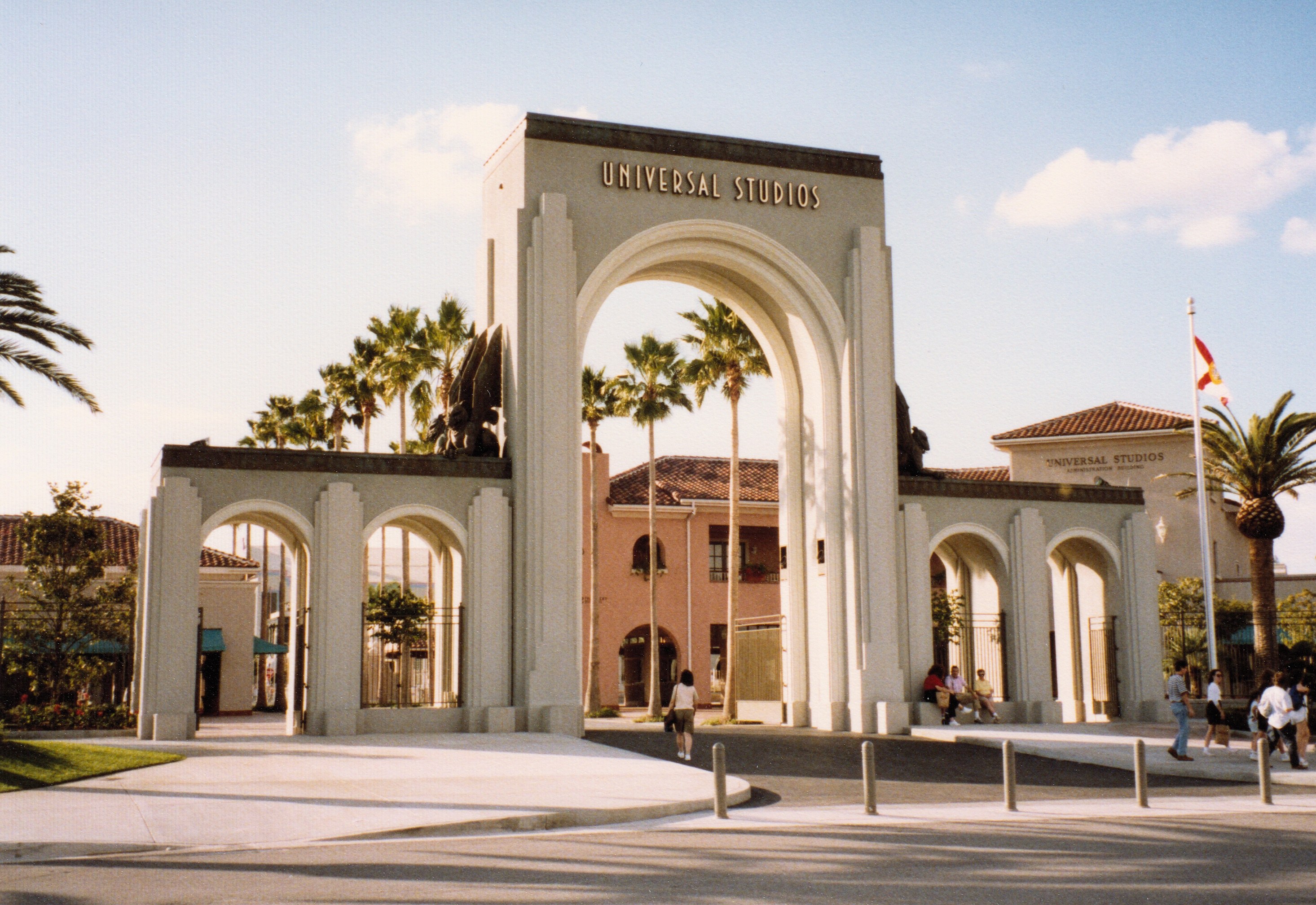 Eingang zum Universal Studios Hollywood mit einem Bogen, einer Statue, Menschen, Bäumen, Gebäuden, einem Fahnenmast und einem bewölkten Himmel.