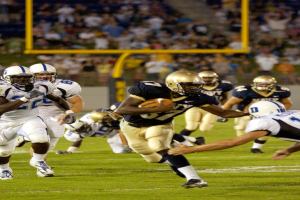 Ein Füßballspieler in einem Sportoutfit und Helm rennt mit dem Ball, umgeben von anderen Spielern, mit unscharfen Hintergrundelementen und Text unten.