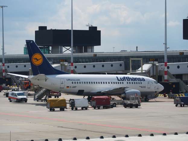 Ein Lufthansa Airbus A320-200 am Frankfurter Flughafen mit Bodenfahrzeugen und Wagen davor, vor einem Gebäude, Laternenmasten und einem klaren blauen Himmel.