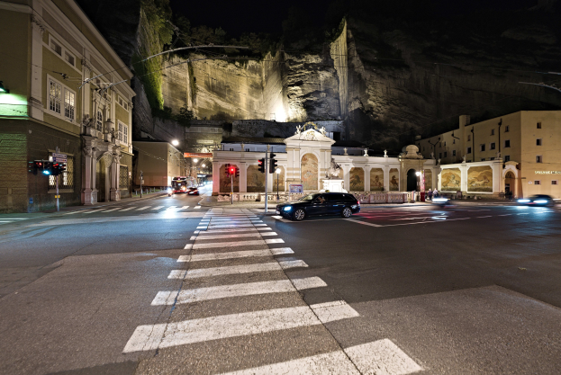 Eine nächtliche Stadtstraße mit fahrenden Autos, Straßenlaternen, Ampeln, beleuchteten Gebäuden, Laternenpfählen, einem Hügel im Hintergrund und einer Zebrastreifenüberquerung unten.