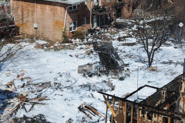 Die Überreste eines durch einen Brand zerstörten Hauses in einer verschneiten Wohngegend, mit Bäumen und Gebäuden im Hintergrund.