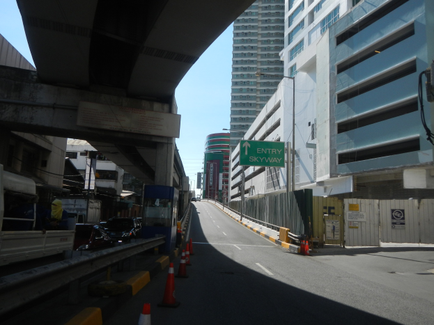 Stadtstraße mit Verkehrskegeln, Fahrzeugen, einer Brücke, Gebäuden mit Namenschildern, Laternenmästen und einem Himmel als Hintergrund.