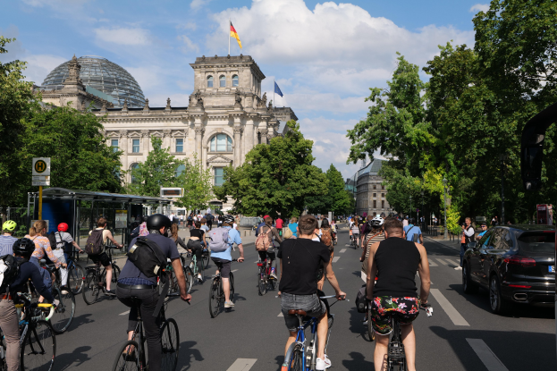 Eine Gruppe von Menschen, die auf Fahrrädern eine von Bäumen gesäumte Straße in Berlin, Deutschland, entlangfahren, mit einem Bushaltestellen auf der rechten Seite und einer Flagge, die auf einem Gebäude weht, unter einem bewölkten Himmel.