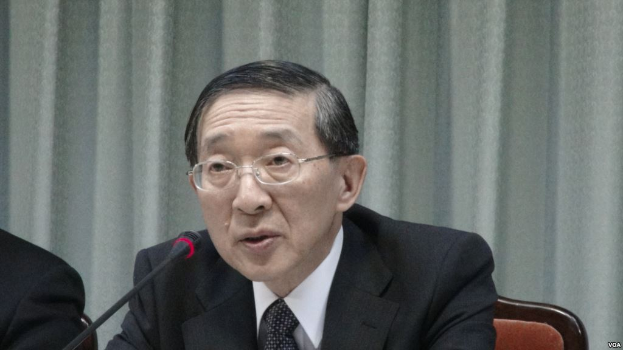 Ein Mann im Anzug und Krawatte sitzt an einem Tisch mit einem Mikrofon, trägt eine Brille und ist in einem formellen Setting mit einem Vorhang im Hintergrund zu sehen, während die Hand einer anderen Person auf der linken Seite des Bildes zu erkennen ist.