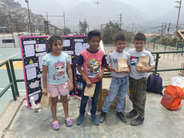 Gruppe von Kindern auf einem Basketballfeld mit Papiertüten in den Händen, Tüten und Gegenstände rechts und eine Tafel mit Text links, vor einem Hintergrund aus Zaun, Pfosten, Straßenlaternen, Bäumen, Gebäuden, Hügeln und Himmel.