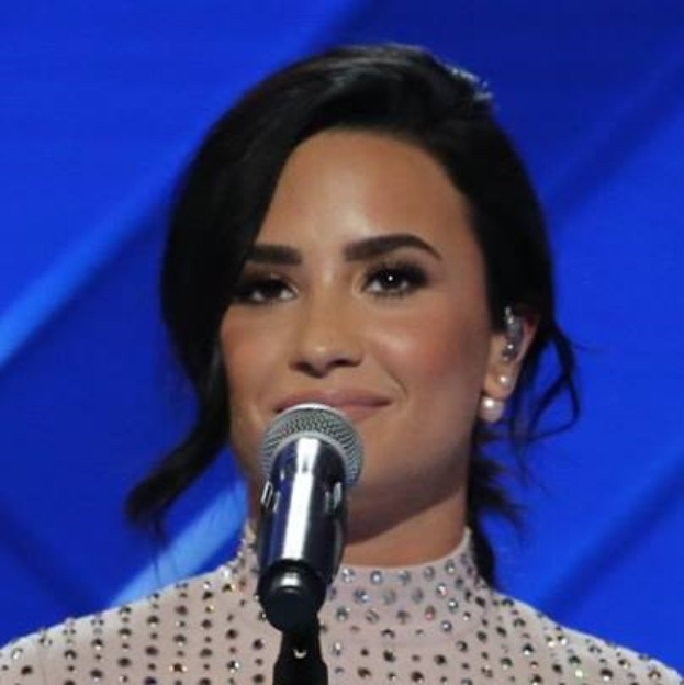 Demi Lovato steht mit einem Mikrofon auf der Bühne bei den Grammys, trägt ein schwarzes Kleid und eine glatte Bob-Frisur, mit einem blauen Hintergrund dahinter.