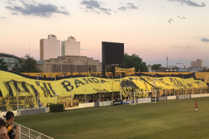 Ein Fußballfeld mit Menschen darauf, umgeben von einem Metallzaun, einem Torpfosten, einer Banner mit der Aufschrift "Boca Juniors vs Real Sociedad", Pflanzen, einem Anzeigebildschirm, Pfählen, Baumrinde, Bäumen, Gebäuden und einem bewölkten Himmel.