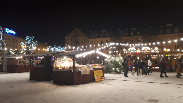 Ein verschneiter Weihnachtsmarkt bei Nacht mit Menschen, Buden, Pflanzen, Gebäuden und Schildern unter einem bewölkten Himmel.