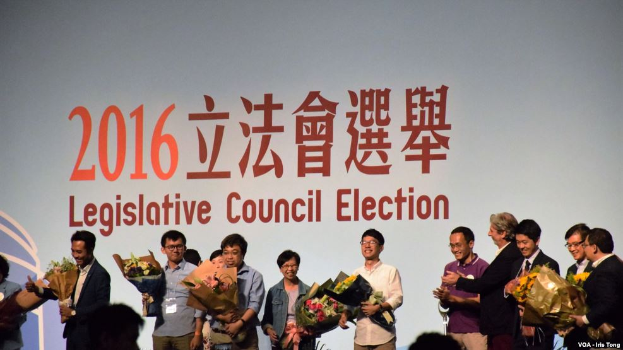 Eine Gruppe von Menschen auf einer Bühne mit Blumensträußen, mit einem Publikum darunter und einem Bildschirm mit Text und einem Logo für die Wahl des Legislativrats im Hintergrund.