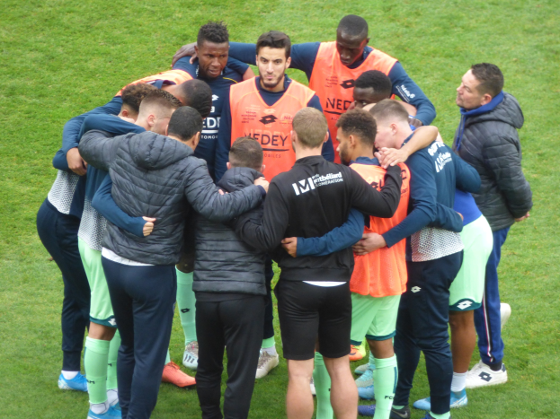 Eine Gruppe von Männern in Jacken, die sich im Kreis auf einem Fussballfeld zusammendrängen.