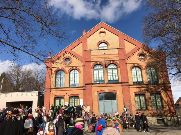 Eine große Gruppe von Menschen steht vor einem mehrstöckigen Gebäude mit Fenstern und Türen, umgeben von Bäumen unter einem bewölkten Himmel, wahrscheinlich bei einem Stadtfest.
