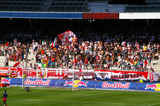 Ein Fußballspiel in einem Stadion mit Spielern auf dem Feld, ein Tor mit Netz, Banner, ein Metallzaun, eine Anzeigetafel, ein Display und ein Dach mit Deckenleuchten.