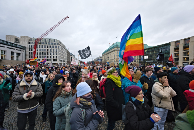 Eine große Gruppe von Menschen mit LGBTQ+-Rechten-Bannern und -Fahnen vor einem Berliner Gebäude, mit einem Kran und einem bewölkten Himmel im Hintergrund.