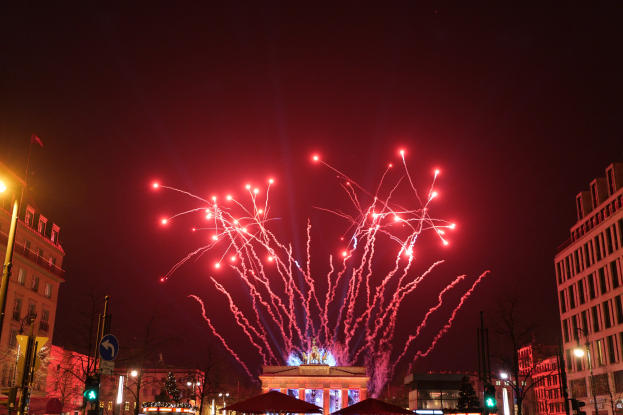Eine Straßenzeile an Silvester in Berlin, voller Gebäude, Bäume, Laternenpfähle, Verkehrsampeln, Schilder, Zelte und Menschen, mit einem festlichen Feuerwerk, das den Nachthimmel erhellt.