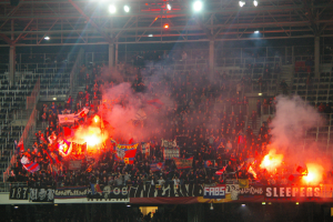 Große Menschenmenge in einem Stadion mit Fackeln und Rauch, haltend Fahnen und Banner, unter einem Dach mit Deckenleuchten und Metallrahmen.