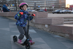 Ein junger Junge fährt mit einem Helm und Handschuhen auf einem Gehweg, mit verschiedenen städtischen Elementen und einem klaren blauen Himmel im Hintergrund.