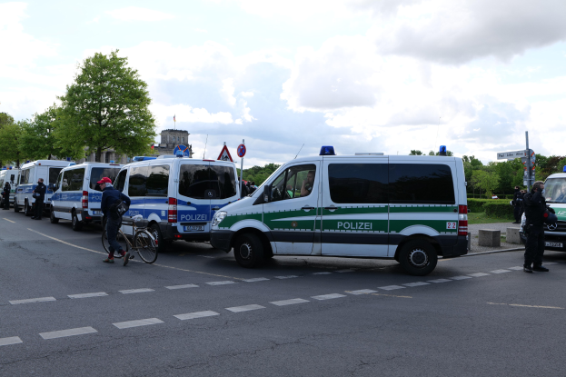 Eine Gruppe von Polizeiwagen entlang einer Straße geparkt mit einem Radfahrer im Vordergrund, Bäume und Gebäude im Hintergrund unter einem bewölkten Himmel.