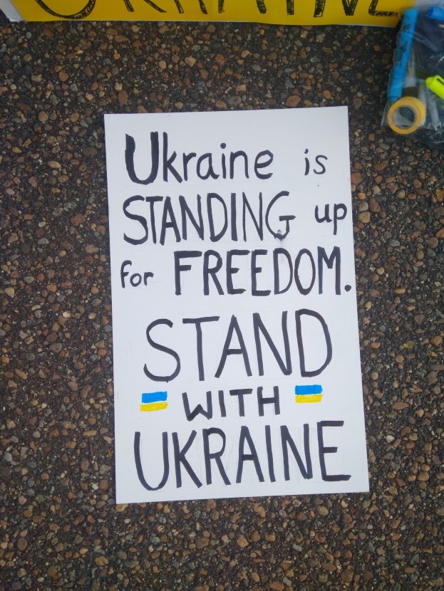 Weißer Papierbogen mit dem Text "Ukraine steht für Freiheit ein, stellt Euch mit Ukraine" auf dem Boden vor einer gelben Fahne, mit einer Plastikhülle mit verschiedenen Gegenständen rechts daneben.