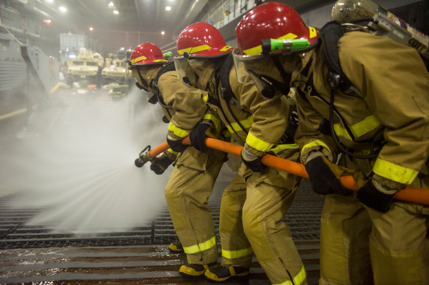 Eine Gruppe von Feuerwehrleuten in Helmen und Handschuhen hält Schläuche und sprüht Wasser auf ein Feuerwehrauto, mit verschiedenen Gegenständen und Texttafeln im Hintergrund und einem Boden unten.
