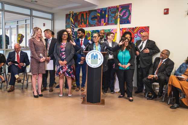 Eine Gruppe von Menschen, die vor einem Podium mit Mikrofon und einer Tafel mit der Aufschrift "Sancho City Councilman Joins City Council" steht und sitzt, mit Flaggen, Feuerlöscher und Deckenlampen im Hintergrund.