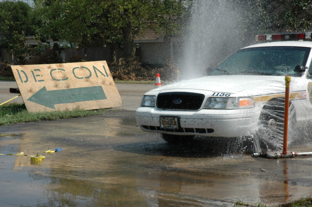 Ein Streifenwagen fährt durch eine Pfütze, mit einem Schild, einer Verkehrsinsel und anderen Gegenständen auf dem Boden, vor einem Hintergrund aus Bäumen, Gebäuden und einem klaren blauen Himmel.