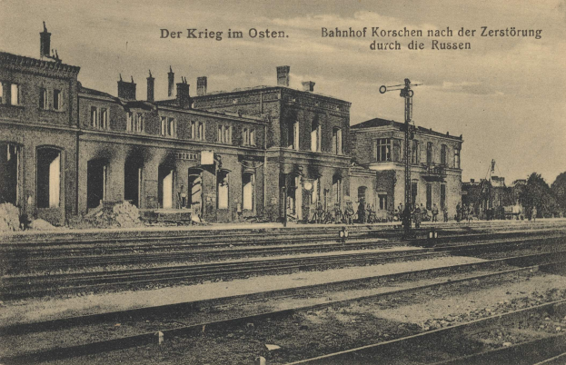 Altes Schwarz-Weiß-Foto vom Bahnhof Korschen in Ostern, Deutschland, das eine Bahnstation mit Gleisen, Gebäuden, Pfählen, Menschen, Bäumen und einem Himmel im Hintergrund zeigt.