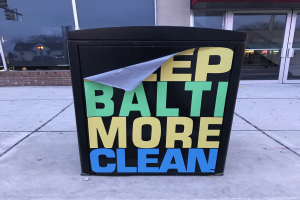 Ein schwarzer Mülltonnen mit "Keep Baltimore More Clean" darauf, vor einem Gebäude mit Glastüren auf dem Bürgersteig platziert.