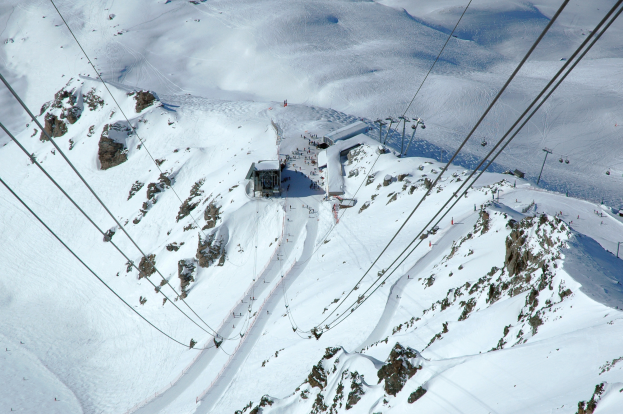 Eine Luftaufnahme eines Skilifts, der einen schneebedeckten Berg hinabfährt, mit sichtbaren Seilbahnkabeln, Felsen und Häusern bei strahlendem Sonnenschein.
