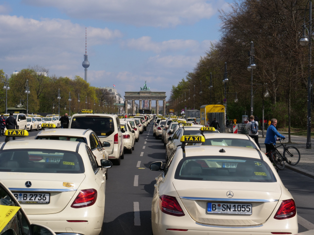 Eine lange Reihe von Taxis steht entlang einer belebten Straße in Berlin, Deutschland, mit Fahrrädern und Fußgängern auf dem Gehweg, flankiert von Bäumen und Laternenpfählen und Gebäuden, einem Bogen und einem Turm im Hintergrund unter einem bewölkten Himmel.
