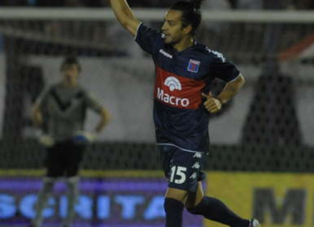 Ein Fußballspieler feiert nach einem Tor auf dem Boden, mit jubelnden Zuschauern und einer Person im Hintergrund und einer Tafel mit Text dahinter, alles vor einem unscharfen, dynamischen Hintergrund.