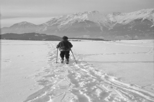 Eine Person Skilanglauf im Schnee mit Bergen im Hintergrund, trägt eine Jacke, Mütze und Handschuhe und hält Ski-Stöcke in den Händen.