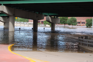 Eine überflutete Stadtstraße unter einer Brücke, mit Wasser bis zur Brückenhöhe, einem Geländer auf der rechten Seite und Bäumen und Gebäuden im Hintergrund.