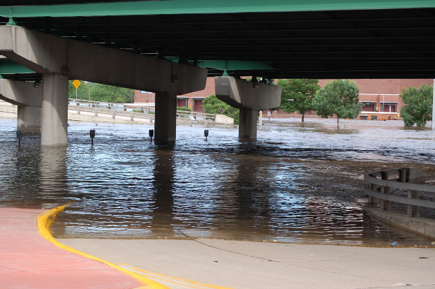 Eine überflutete Stadtstraße unter einer Brücke, mit Wasser bis zur Brückenhöhe, einem Geländer auf der rechten Seite und Bäumen und Gebäuden im Hintergrund.