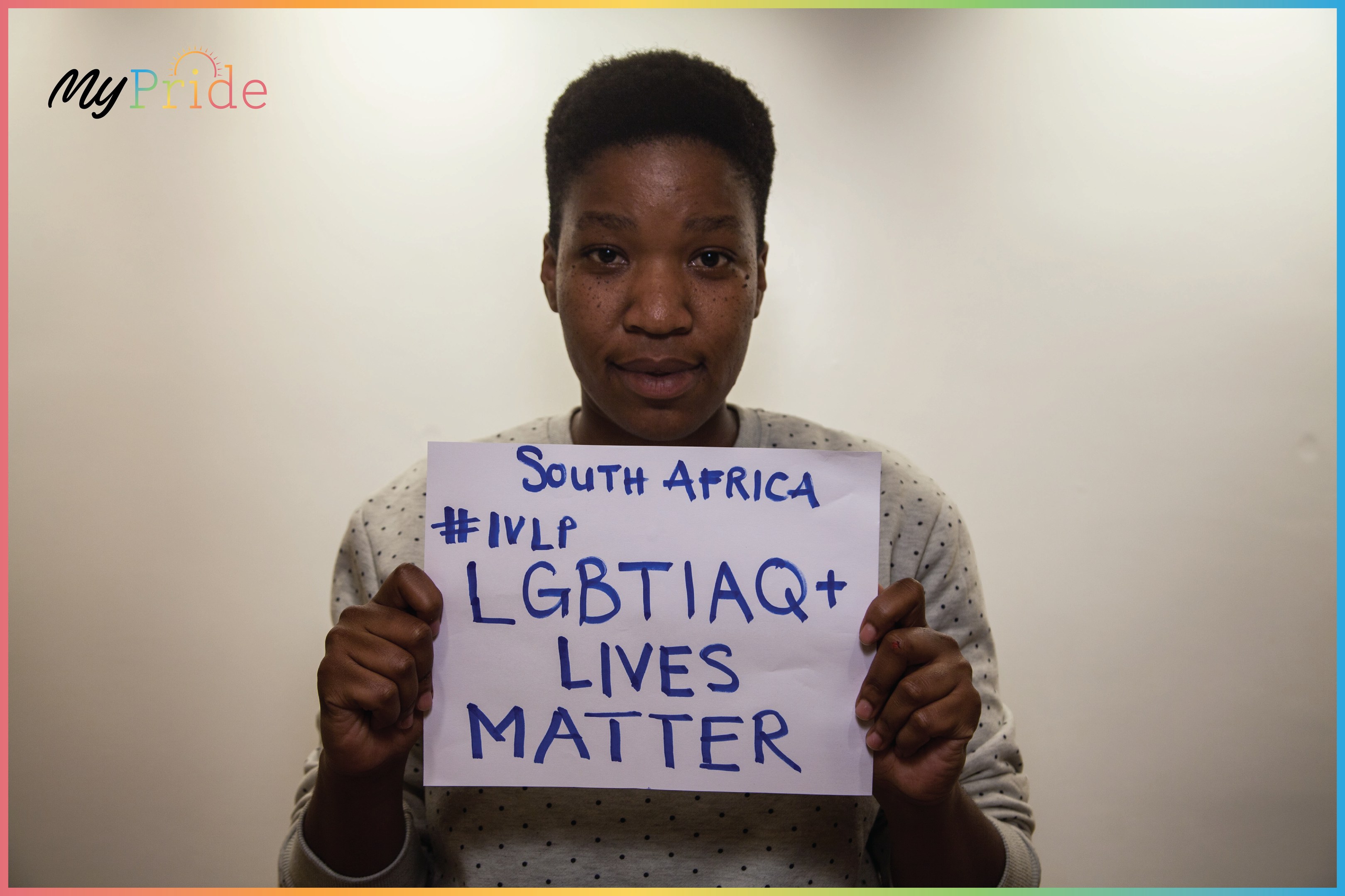 Frau in weißem Hemd mit einem Schild, auf dem "LGBTQ+ Lives Matter" steht, vor einer Wand mit entschlossener Miene.