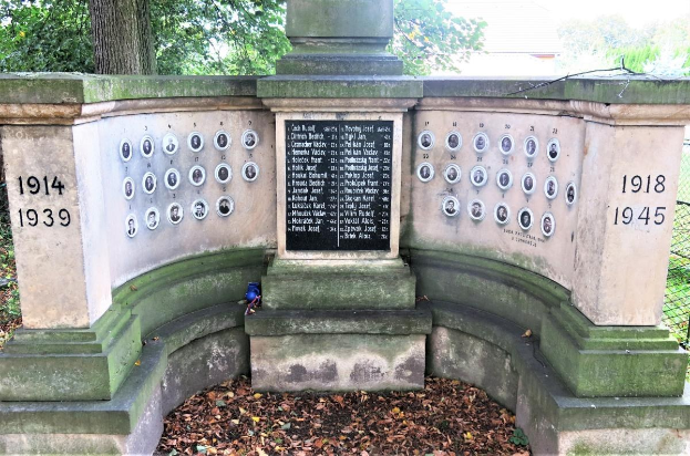 Ein Holocaust-Gedenkmonument in einem jüdischen Friedhof in Berlin, mit einer Text- und Nummerntafel an seiner Wand, umgeben von Bäumen, einem Zaun und trockenen Blättern auf dem Boden.