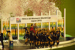 Eine Gruppe von Fussballspielerinnen in schwarzen Uniformen feiert ihren Sieg auf einer Bühne, umgeben von einer Menge mit Kameras, mit einem dekorierten Hintergrund und grasbewachsenem Boden.