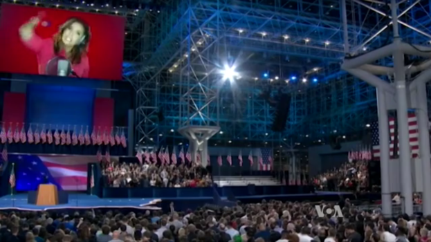 Eine große Menschenmenge sitzt vor einer Bühne mit einem Rednerpult, Mikrofon, Flaggen, einem Bildschirm mit einer Frau und Lichtausrüstung, wahrscheinlich bei einer Präsidentschaftsdebatte.