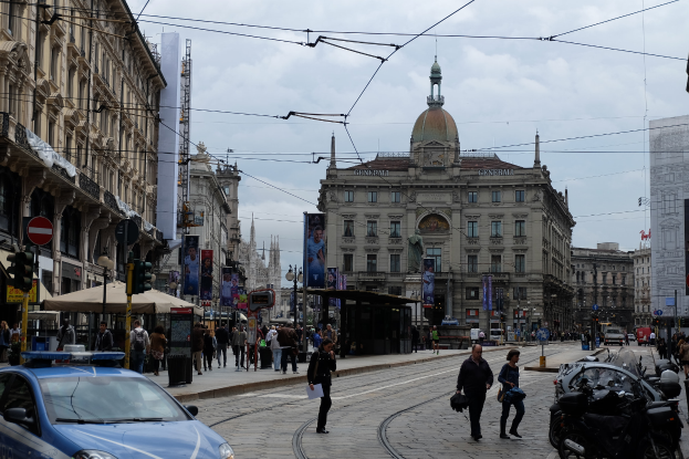 Eine belebte Stadtstraße mit einem parkenden Polizeiauto, Fußgänger mit Taschen, fahrende Fahrzeuge, Gebäude mit Fenstern, Banner, Laternenpfähle, Verkehrsampeln und ein bewölkter Himmel.