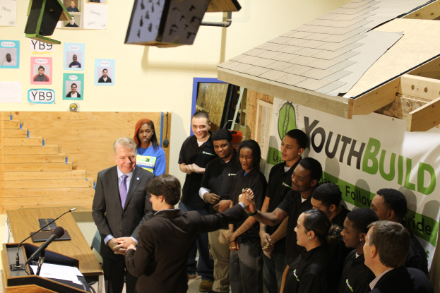 Gruppe von Menschen, die sich die Hände schütteln, um einen Podium mit Mikrofonen und Papieren auf einem Tisch, vor einem Publikum, mit Plakaten an der Wand und Gegenständen auf dem Dach.
