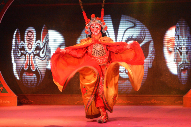 Eine Frau in einem roten Kleid führt eine Chinesische Oper auf der Bühne auf, trägt ein buntes Kostüm und bewegt sich anmutig zur Musik, mit einem Sichtschutz im Hintergrund.