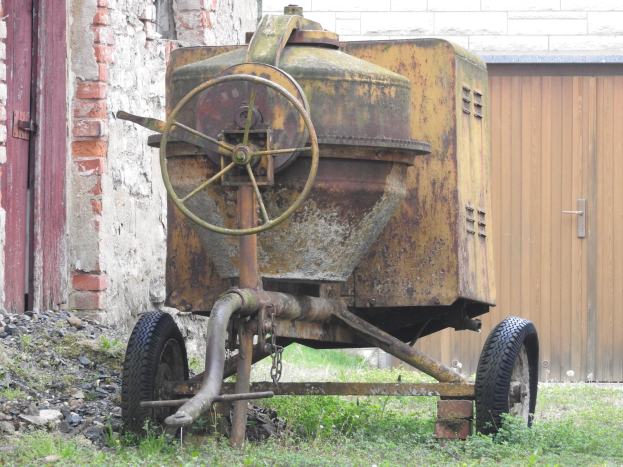Alter rostiger Anhänger vor einem Backsteingebäude geparkt, mit Gras und Steinen drumherum und einer Wand und einer hölzernen Tür im Hintergrund.