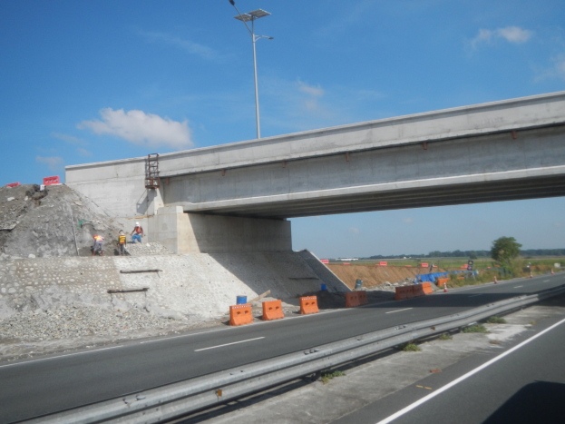 Autobahn mit Brücke darüber, ein Auto darunter vorbeifahrend, Bauarbeiter am Straßenrand, Verkehrskegel, Steine und eine Straßenlaterne, mit Bäumen und einem bewölkten Himmel im Hintergrund.