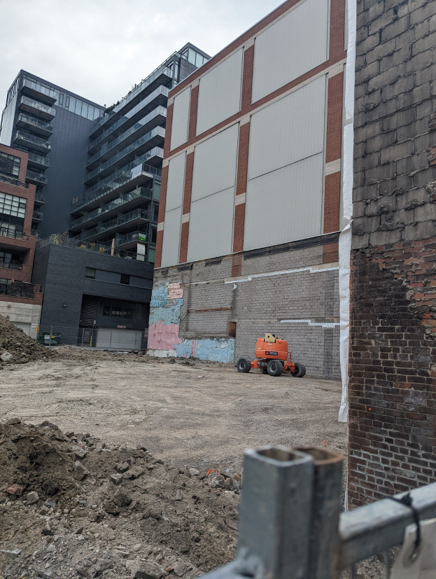Baustelle mit einem teilweise abgerissenen Gebäude im Hintergrund, einem Fahrzeug auf dem Boden, einem Metallzaun und einem bewölkten Himmel.