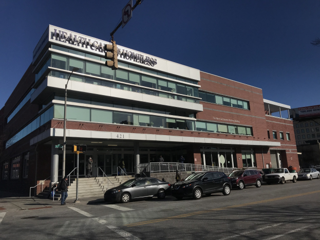 Großes Gebäude mit der Aufschrift "New York City Health Care Homeless Center" mit parkenden Autos, Passanten, Straßenmöbeln und Bäumen unter einem klaren Himmel.