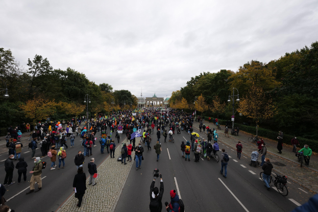 Eine große Gruppe von Menschen marschiert auf einer von Bäumen gesäumten Straße mit Laternenmasten, die Kameras halten, mit einem Gebäude und einem klaren Himmel im Hintergrund, was auf eine Demonstration in Berlin hindeutet.